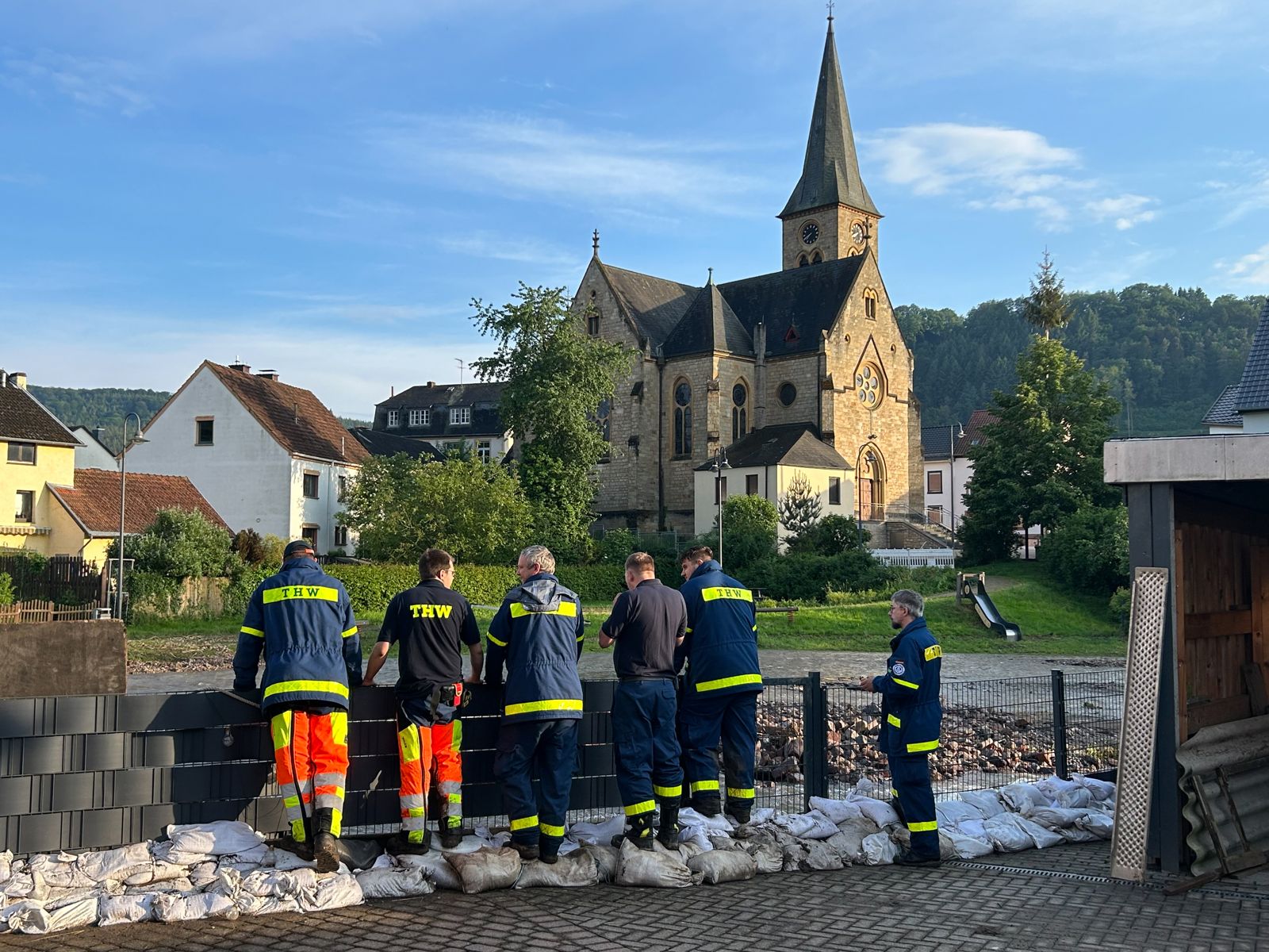THW-Helfer und Feuerwehr sichern mit einer Sandsackreihe das Ufer eines Hochwasser führenden Bachs, im Hintergrund eine Dorfkirche