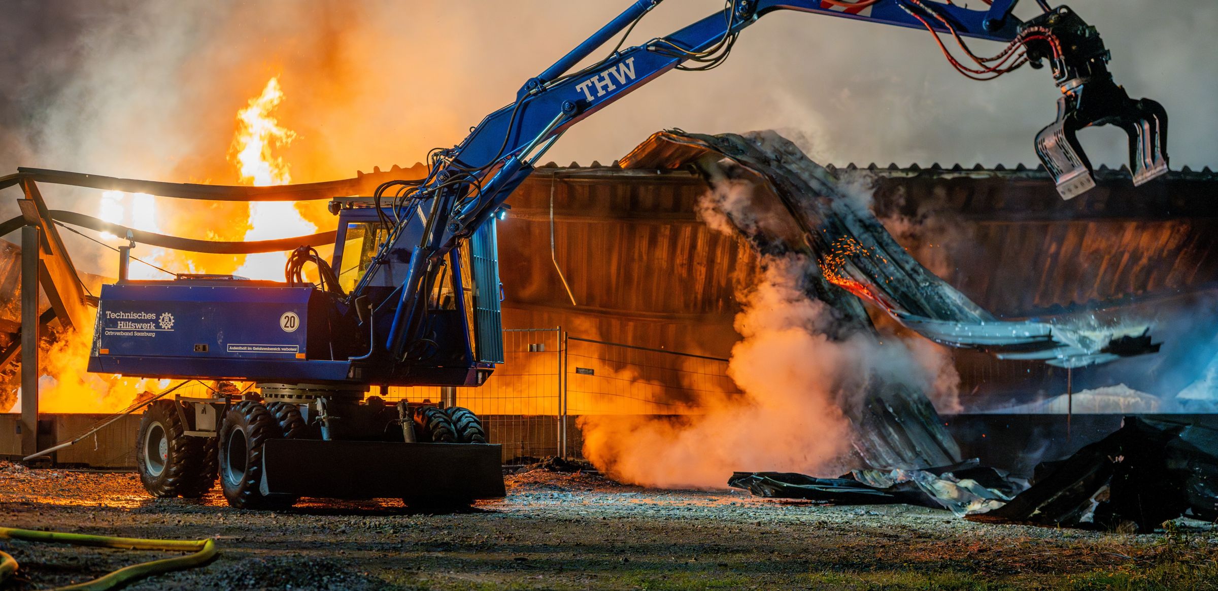 THW-Bagger räumt bei einem nächtlichen Großbrand Trümmer, im Hintergrund lodern Flammen